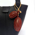 A black textured leather tote bag featuring a decorative hanging charm. The charm consists of two brown leather coffee beans attached to the handle with a light tan cord.