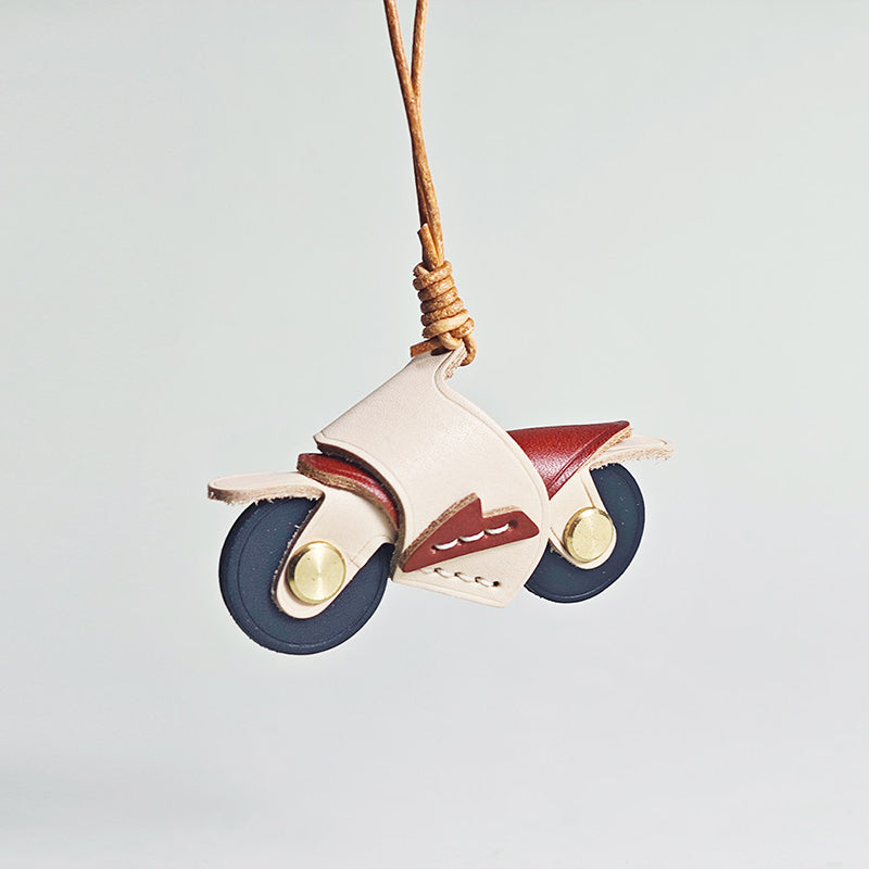 A hanging view of a handcrafted leather motorcycle charm. The body is cream-colored with a dark red seat and navy blue wheels, suspended by a twisted leather strap.