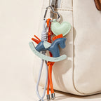 A lifestyle shot showing the blue and mint green leather rocking horse keychain attached to the zipper of a beige canvas handbag. The colorful charms and braided cords add a playful accent to the accessory.