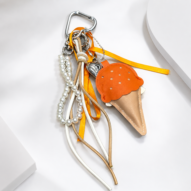 A close-up view of an orange leather ice cream cone bag charm with a white scalloped edge and perforated details, attached to a silver carabiner clip. It features orange and white ribbons, a pearl chain, and a small silver heart charm, highlighting the detailed craftsmanship of this trendy accessory.