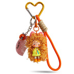A gold-tone heart clip keychain holding a doll with bright orange curly hair and a lime green top. The accessory comes with a matching orange rope lanyard and a miniature comb pendant.