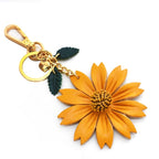 A side-angle shot of an orange leather daisy keychain against a white background. The charm includes a gold-toned clip, a circular ring, and two textured green leaves dangling beside the bloom.