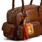 A brown leather handbag accessorized with the orange curly-haired doll keychain. The charm is clipped onto the bag's handle hardware, showing how it looks when worn as a bag pendant.