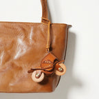 An orange leather motorcycle keychain attached to the handle of a brown leather tote bag. The charm hangs naturally, showcasing its use as a stylish bag accessory.