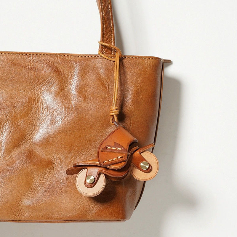An orange leather motorcycle keychain attached to the handle of a brown leather tote bag. The charm hangs naturally, showcasing its use as a stylish bag accessory.