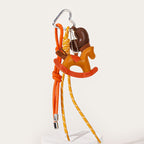 A close-up of a handmade leather keychain featuring an orange rocking horse charm. It is attached to a silver heart-shaped carabiner with orange and yellow braided cords hanging down.