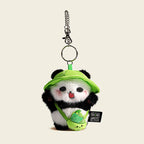 A plush panda bag charm wearing a green bucket hat and carrying a small green bag shaped like a bamboo shoot. The panda has black eyes, a red tongue, and pink cheeks, attached to a silver lobster clasp and keyring, designed as a cute and trendy accessory for bags or keychains.