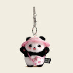 A plush panda bag charm wearing a pink bucket hat and carrying a small pink bag shaped like a flower. The panda has black eyes, a red tongue, and pink cheeks, attached to a silver lobster clasp and keyring, designed as a cute and trendy accessory for bags or keychains.