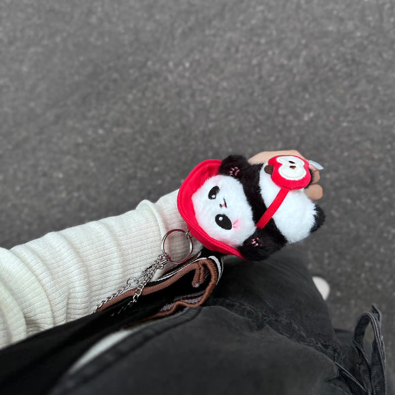 A person wearing a white long-sleeve shirt holds a plush panda bag charm in red, featuring a red hat and a small red bag shaped like an apple. The charm is attached to a silver keyring and hangs from a black bag, adding a playful touch to the outfit.