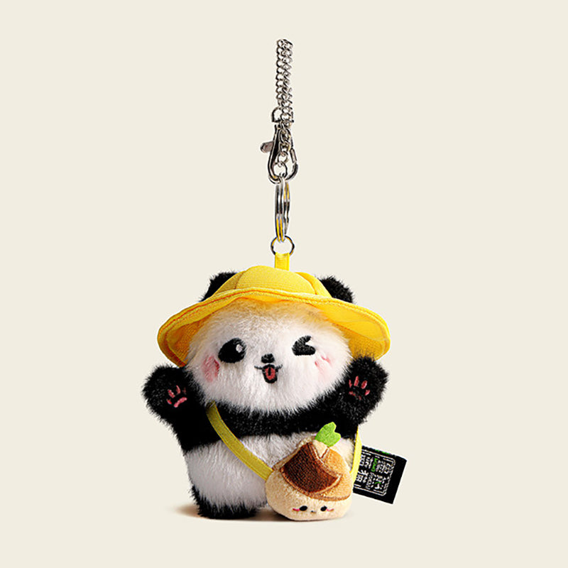 A plush panda bag charm wearing a yellow bucket hat and carrying a small yellow bag shaped like a cake. The panda has black eyes, a red tongue, and pink cheeks, attached to a silver lobster clasp and keyring, designed as a cute and trendy accessory for bags or keychains.