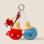 A set of two plush ping-pong paddle keychains, one red and one blue, both with smiling faces and white pom-pom handles, hanging from a keyring with a small acrylic paddle charm.