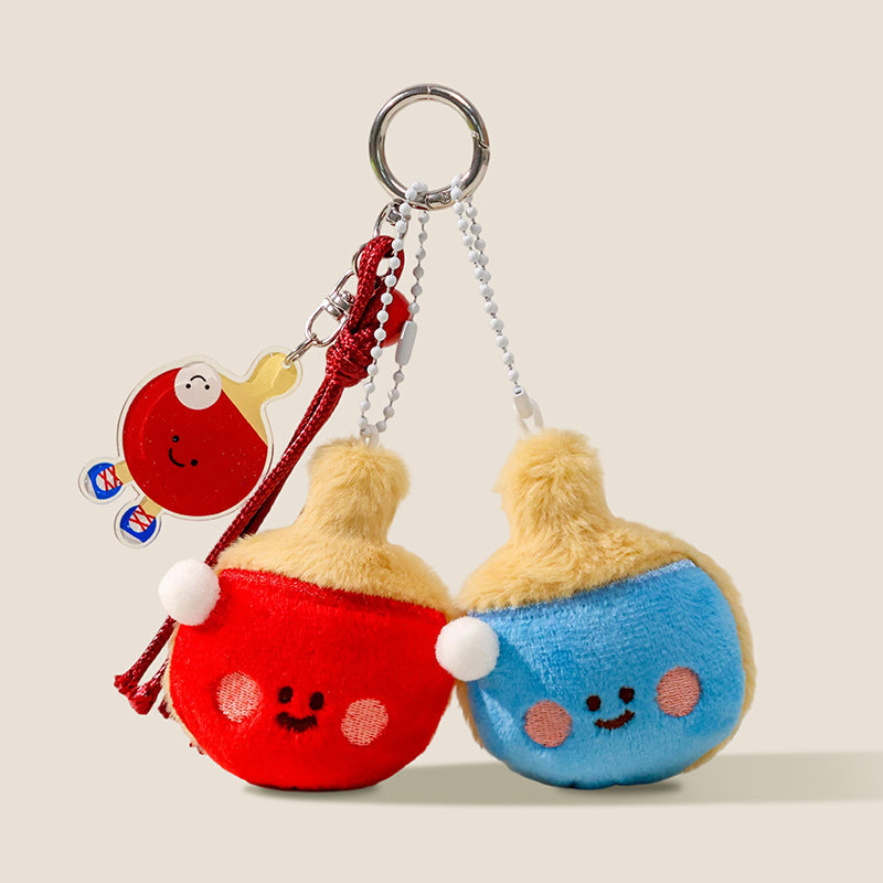 A set of two plush ping-pong paddle keychains, one red and one blue, both with smiling faces and white pom-pom handles, hanging from a keyring with a small acrylic paddle charm.