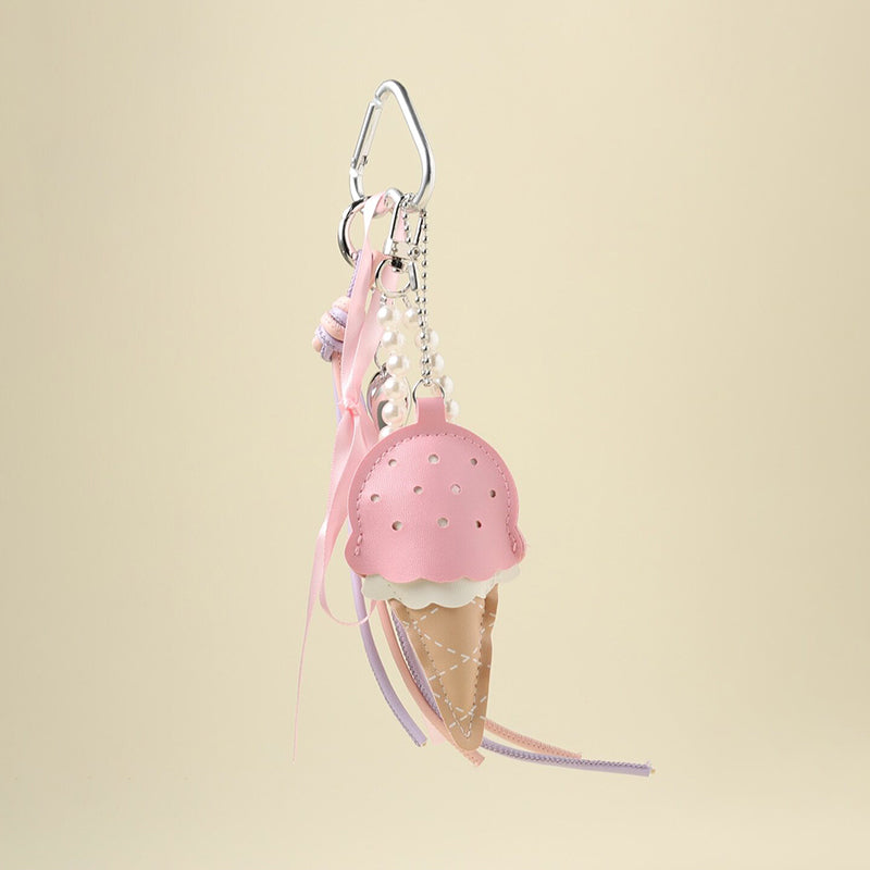 A pink leather ice cream cone bag charm with a white scalloped edge and perforated details, attached to a silver carabiner clip. It features pink and purple ribbons, a pearl chain, and a small silver heart charm, designed as a cute and trendy accessory for bags or keychains.