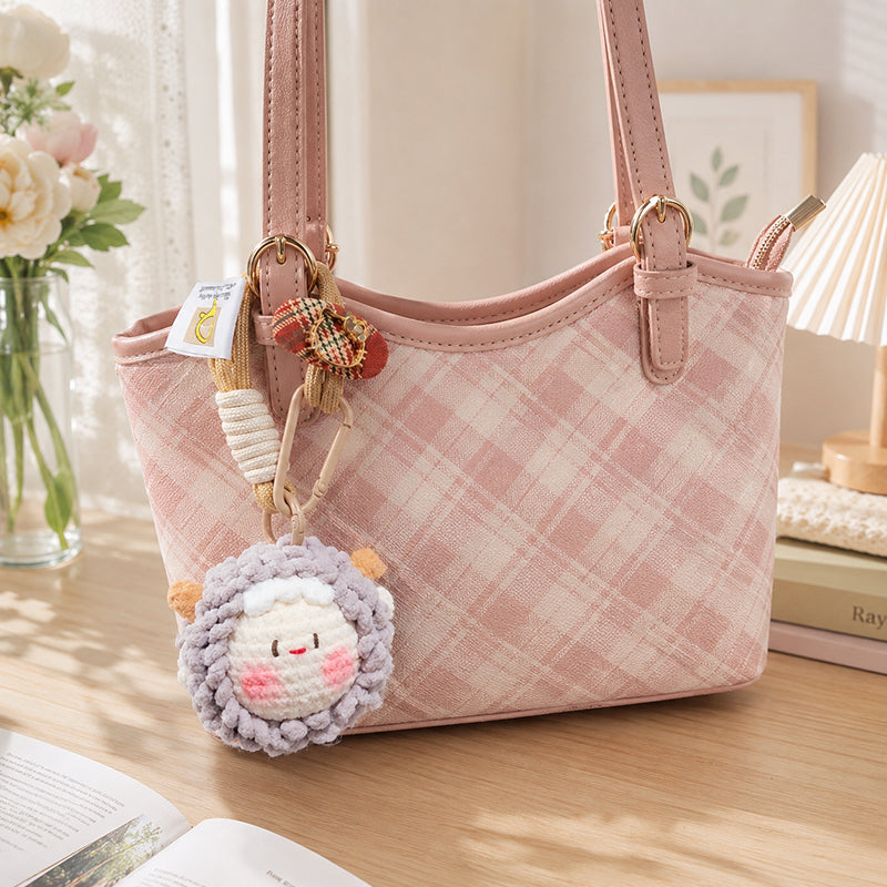 A pink plaid tote bag decorated with a purple crocheted sheep keychain and a red plaid bow charm, placed on a wooden table with books and flowers in the background.