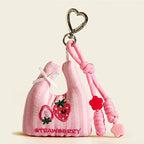 A pink striped fabric bag charm shaped like a mini tote, featuring embroidered strawberries and the word "STRAWBERRY", with a white bow and red flower beads on the strap.