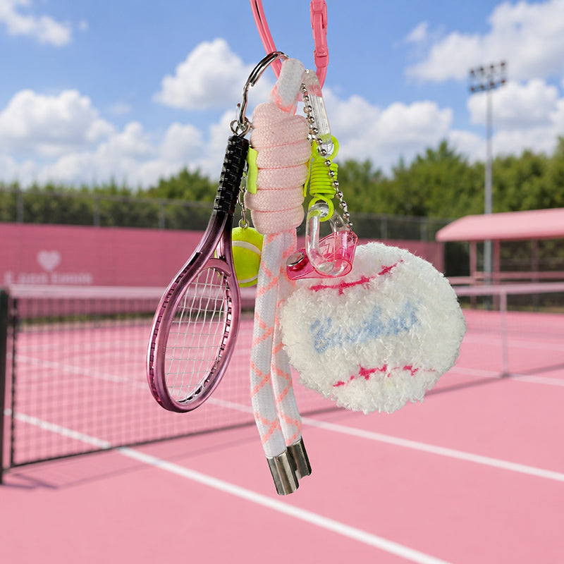 A pink tennis-themed keychain set, including a plush white tennis ball with pink stitching and a miniature pink racket, hangs against the backdrop of a pink tennis court under a blue sky.