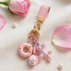 A cute pink-themed keychain set featuring a donut, a Hello Kitty mug, and an iridescent balloon dog. The charms are attached to a pink leather strap with gold hardware, set against a backdrop of pink rose petals and pearls.