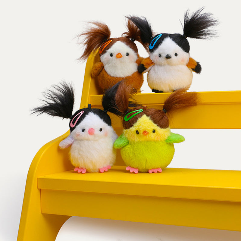Four different colored plush bird keychains (orange, black/white, white/lavender, and lime-green) are displayed together on a bright yellow stepped surface, showcasing their unique color combinations and pigtail hairstyles.

