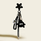 A black, fluffy monster charm with large googly eyes, hanging from a black leather star-shaped strap. It is attached with a black and white striped cord and features a small white tag with a logo.