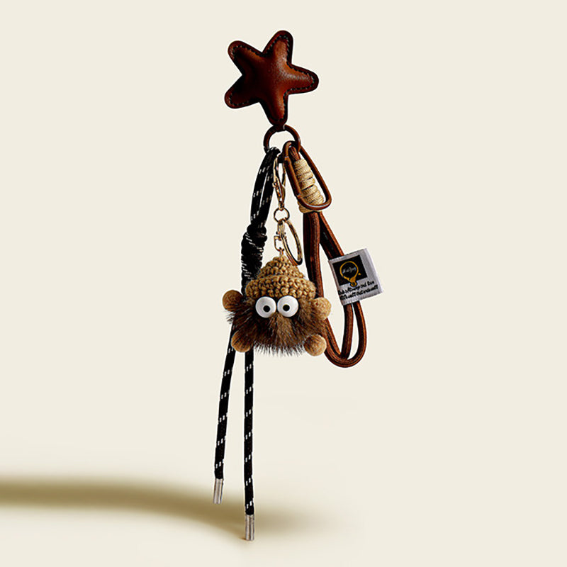 A brown, fluffy monster charm with large googly eyes and a textured, knitted top. It hangs from a brown leather star-shaped strap with a black and white striped cord and a small white tag.
