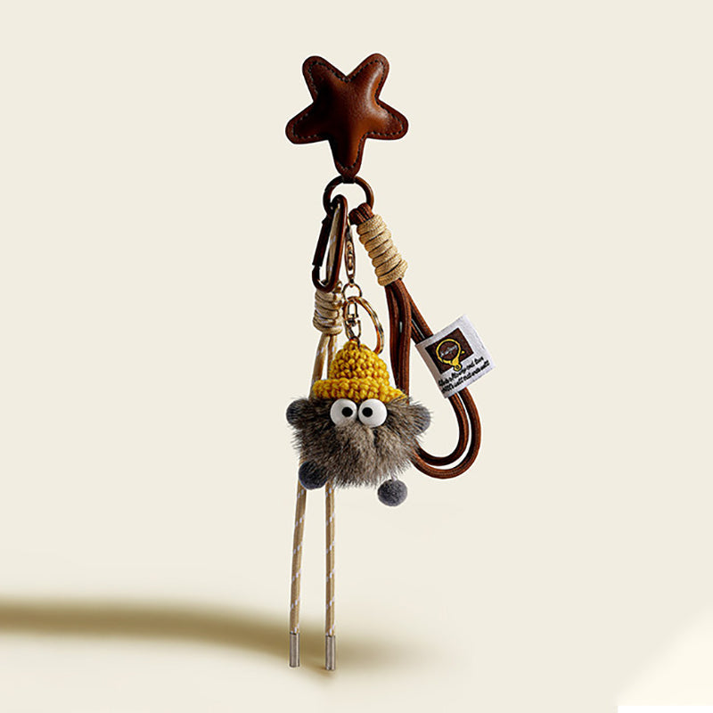 A dark gray, fluffy monster charm with large googly eyes, wearing a yellow knitted hat. It is attached to a brown leather star-shaped strap with a gold-tone clasp and a beige and white striped cord.