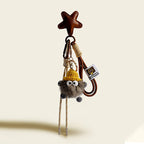 A dark gray, fluffy monster charm with large googly eyes, wearing a yellow knitted hat. It is attached to a brown leather star-shaped strap with a gold-tone clasp and a beige and white striped cord.