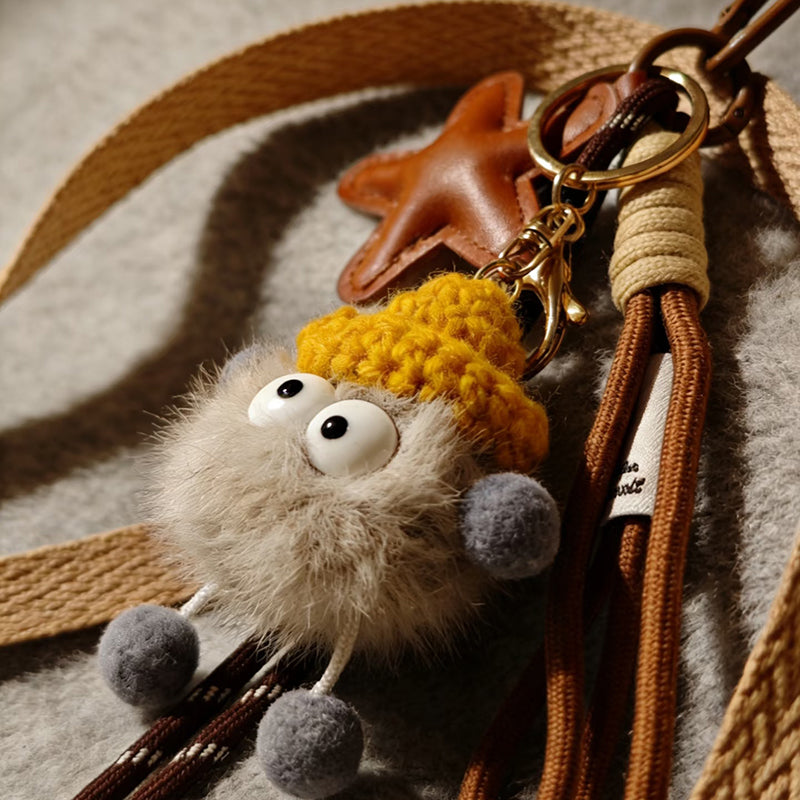 A close-up of a light gray, fluffy monster charm with large googly eyes, wearing a yellow knitted hat. It has small, gray pom-pom feet and is attached to a brown leather strap with a gold-tone clasp, resting on a textured beige surface.