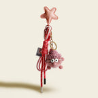 A pink, fluffy monster charm with large googly eyes and a pink knitted hat. It hangs from a pink leather star-shaped strap with a red and white striped cord and a small white tag.