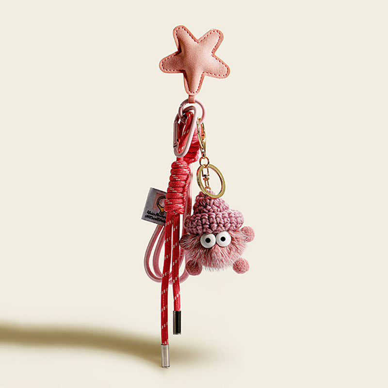 A pink, fluffy monster charm with large googly eyes and a pink knitted hat. It hangs from a pink leather star-shaped strap with a red and white striped cord and a small white tag.