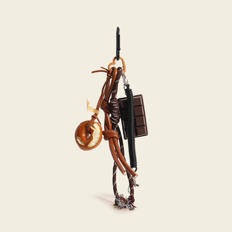 A brown leather bag charm set featuring a donut-shaped charm and a chocolate bar charm, attached to a brown leather strap with a woven rope detail. The charm features a small leather loop and a tag with a logo, designed as a cute and trendy accessory for bags or keychains.