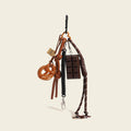 A brown leather bag charm set featuring a pretzel knot-shaped charm and a chocolate bar charm, attached to a brown leather strap with a woven rope detail. The charm features a small leather loop and a tag with a logo, designed as a cute and trendy accessory for bags or keychains.