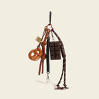 A brown leather bag charm set featuring a pretzel knot-shaped charm and a chocolate bar charm, attached to a brown leather strap with a woven rope detail. The charm features a small leather loop and a tag with a logo, designed as a cute and trendy accessory for bags or keychains.