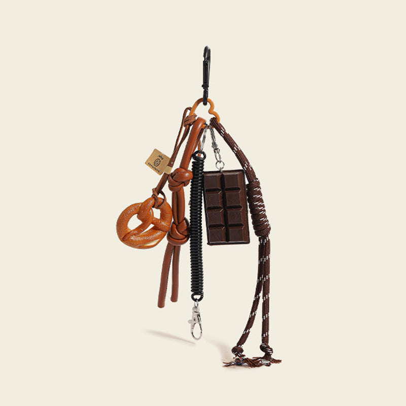 A brown leather bag charm set featuring a pretzel knot-shaped charm and a chocolate bar charm, attached to a brown leather strap with a woven rope detail. The charm features a small leather loop and a tag with a logo, designed as a cute and trendy accessory for bags or keychains.