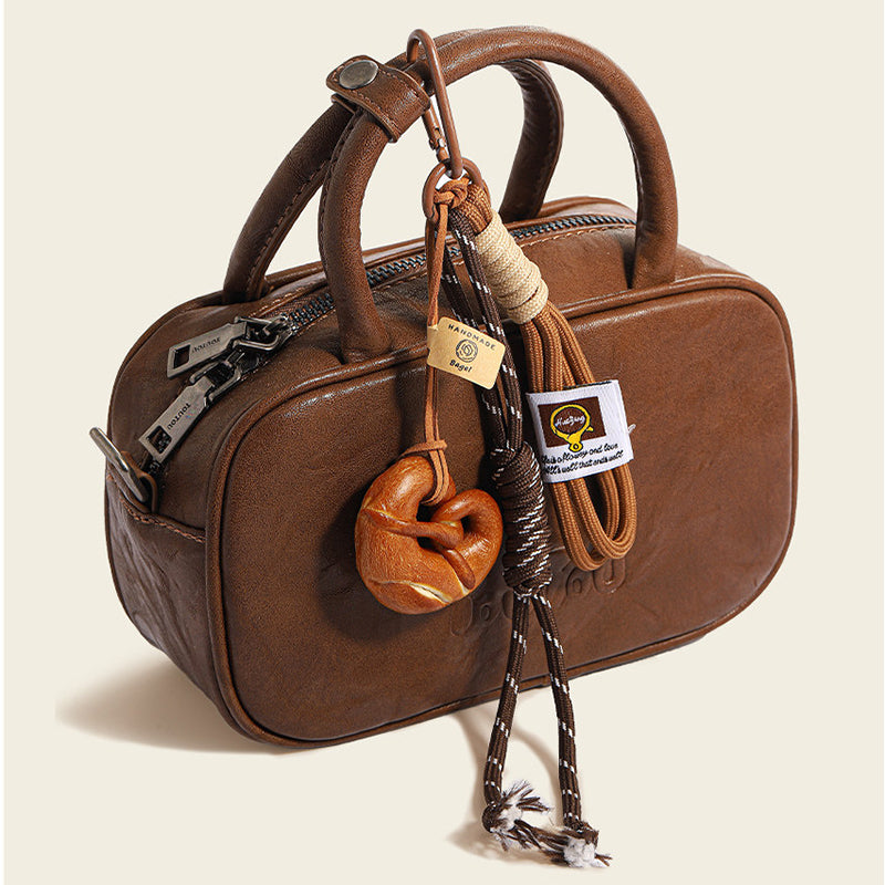 A close-up of a glossy, brown leather handbag featuring a "MEET THE LAURA" embossed logo, with a decorative charm hanging from the strap. The charm includes a realistic-looking pretzel, braided ropes in cream and brown, and a small wooden tag that says "Lucky!".