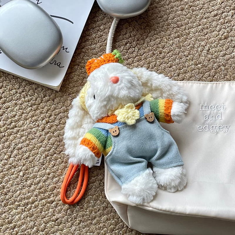 A white plush lop-eared rabbit doll wearing light blue corduroy overalls with bear-shaped buttons and a rainbow-striped knit sweater. It has a crocheted carrot on its head and a yellow flower accessory, lying on a beige carpet next to white headphones and a cream pouch.