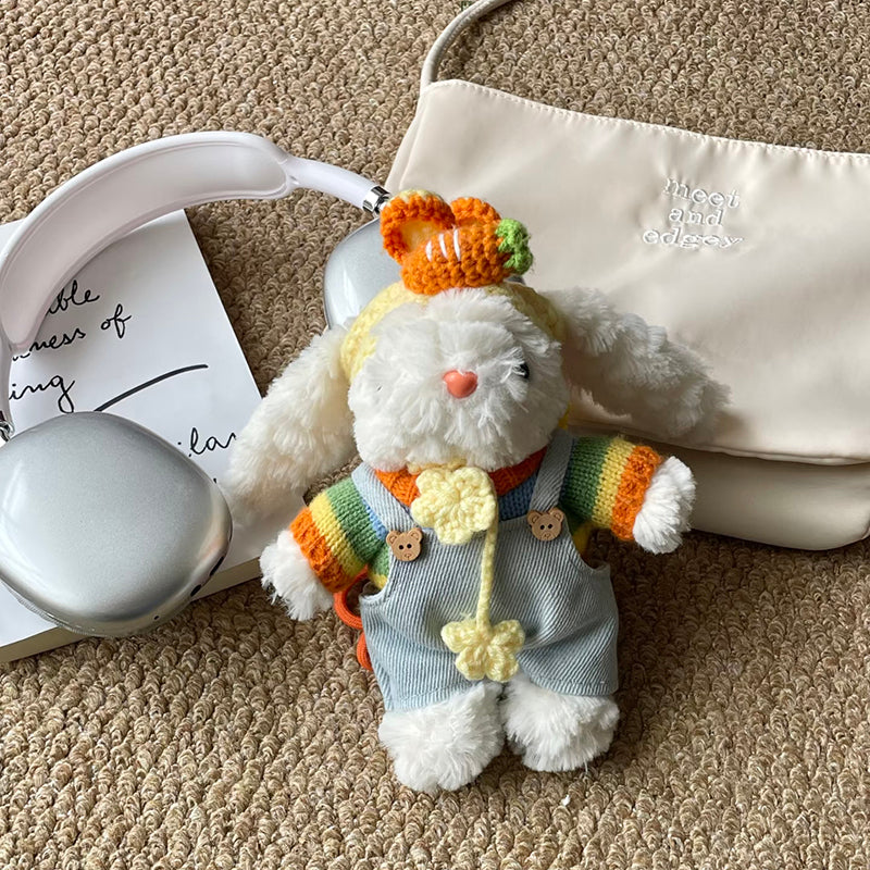 A side view of the white plush lop-eared rabbit doll with a crocheted carrot on its head, wearing rainbow-striped sleeves and light blue overalls. An orange strap is visible, and it is placed next to a cream pouch with "meet and edgey" text and white headphones on a beige carpet.