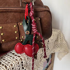 A red leather cherry-shaped bag charm with a dark green stem, hanging from the zipper of a brown leather bag. The charm is attached via a red braided rope strap with a red clip, adding a playful touch to the bag.