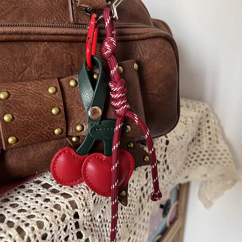 A red leather cherry-shaped bag charm with a dark green stem, hanging from the zipper of a brown leather bag. The charm is attached via a red braided rope strap with a red clip, adding a playful touch to the bag.