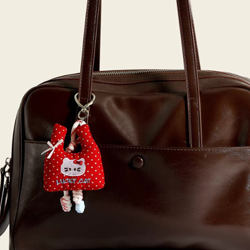 A red polka-dot fabric bag charm shaped like a mini tote, featuring an embroidered "LUCKY CAT" design and a white bow, hanging from a brown leather handbag.