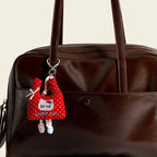 A red polka-dot fabric bag charm shaped like a mini tote, featuring an embroidered "LUCKY CAT" design and a white bow, hanging from a brown leather handbag.