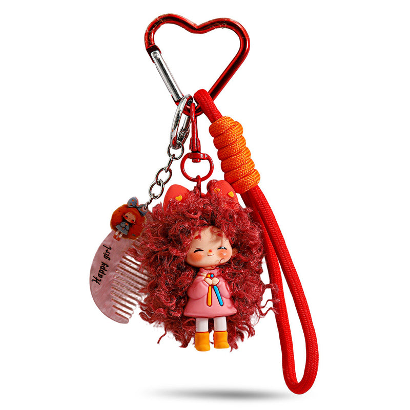 A red heart-shaped carabiner keychain featuring a cute doll with voluminous curly red hair, wearing a pink outfit. It includes a small comb charm inscribed with "Happy girl" and a braided red wrist strap.