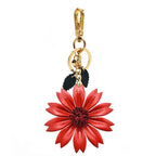 A vibrant red leather daisy pendant hanging vertically from a polished gold-tone carabiner. The flower’s center is detailed with stitched texture, and it is paired with dark green leaf charms.