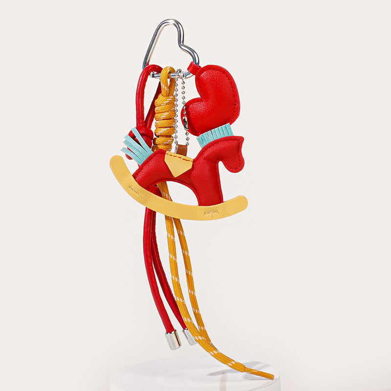 A vibrant red leather rocking horse keychain with a yellow saddle and base. The accessory includes red and yellow braided straps and a small tassel detail, suspended from a metal clip.