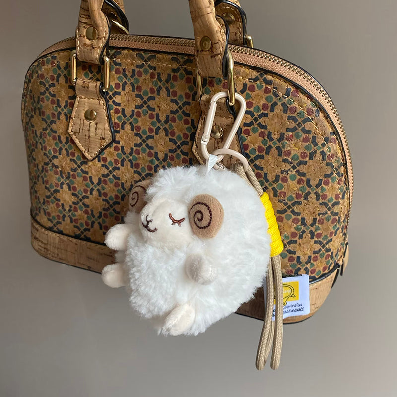 A white fluffy sheep plush charm with closed eyes and brown spiral horns is clipped onto a cork-patterned handbag, with its branded tag visible and hanging down.