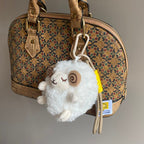 A white fluffy sheep plush charm with closed eyes and brown spiral horns is clipped onto a cork-patterned handbag, with its branded tag visible and hanging down.
