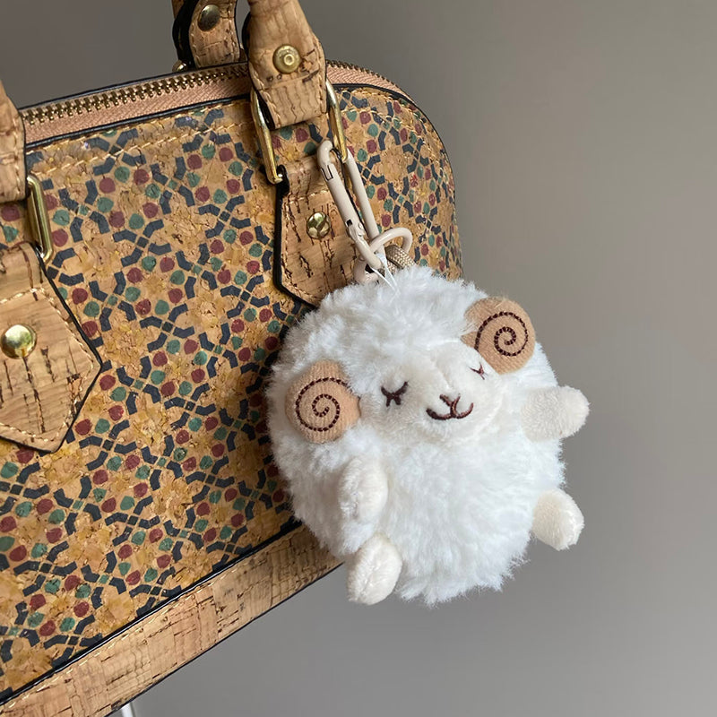 A white fluffy sheep plush charm with closed eyes and brown spiral horns is clipped onto the handle of a cork-patterned handbag with geometric designs.