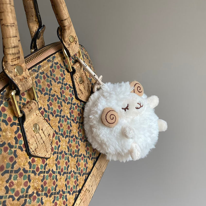 A side-angle shot of a white fluffy sheep plush keychain hanging from a cork-patterned bag, showing its round shape and soft texture.
