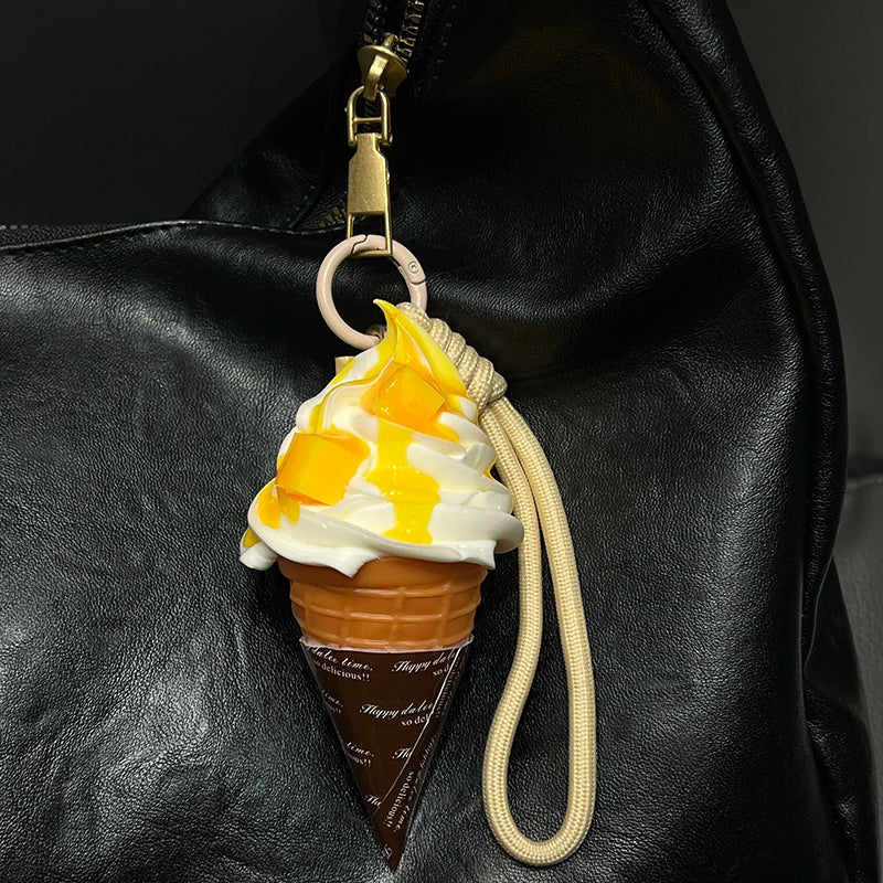 A realistic-looking ice cream cone charm with yellow mango-flavored swirls and white cream, topped with small, fruit-like pieces. It is attached to a beige braided rope with a metal clasp, hanging from the zipper of a black leather bag.