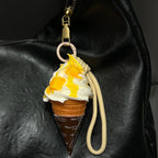 A realistic-looking ice cream cone charm with yellow mango-flavored swirls and white cream, topped with small, fruit-like pieces. It is attached to a beige braided rope with a metal clasp, hanging from the zipper of a black leather bag.