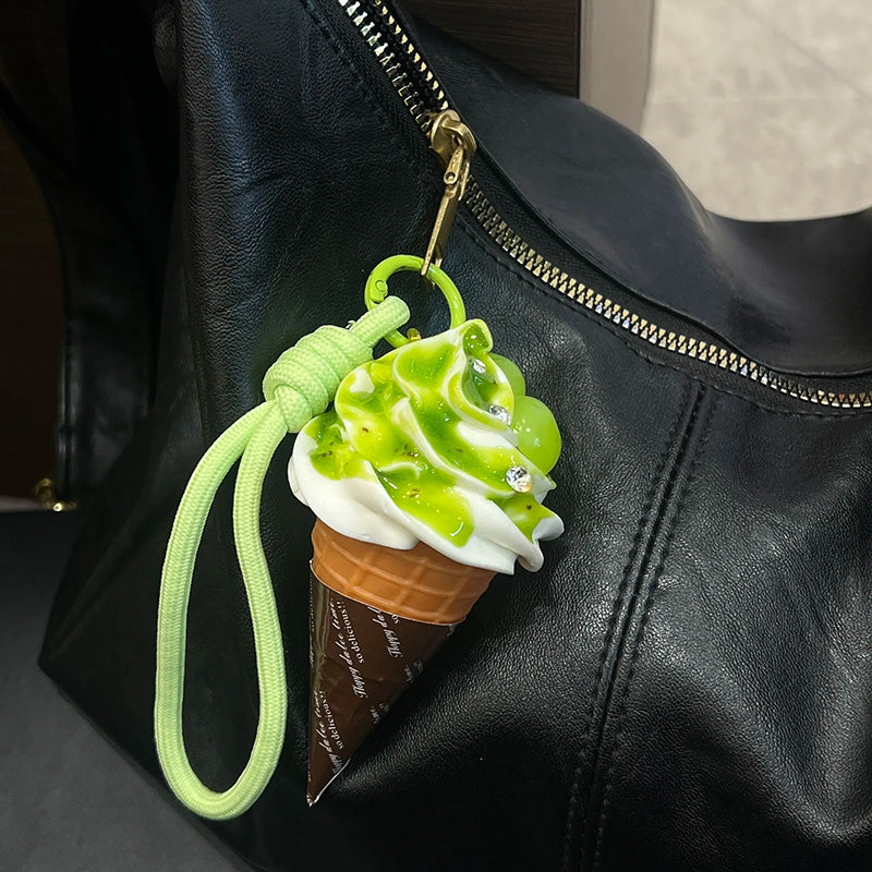 A realistic-looking ice cream cone charm with green matcha-flavored swirls and white cream, topped with small, shiny embellishments. It is attached to a light green braided rope with a metal clasp, hanging from the zipper of a black leather bag.